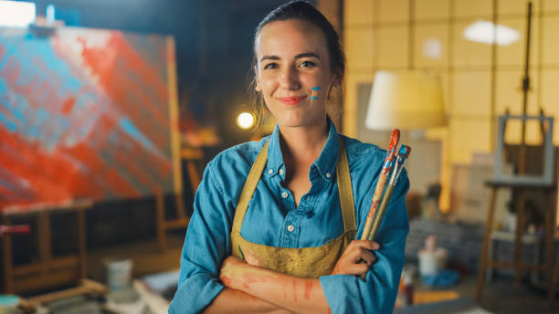 porträt der talentierten jungen künstlerin schmutzig mit farbe, tragen schürze, kreuze arme beim halten pinsel, blick auf die kamera mit einem lächeln. authentisches creative studio mit großem canvas und tools überall - bildender künstler künstler stock-fotos und bilder