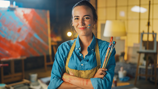 Portrait of Talented Young Female Artist Dirty with Paint, Wearing Apron, Crosses Arms while Holding Brushes, Looks at the Camera with a Smile. Authentic Creative Studio with Large Canvas and Tools Everywhere