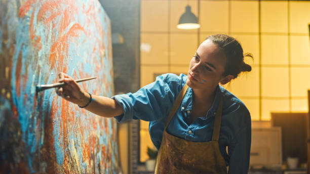 portrait of talented female artist working on a modern abstract oil painting, gesturing with broad strokes using paint brush. dark creative studio large picture stands on easel illuminated. - ocupação artística imagens e fotografias de stock