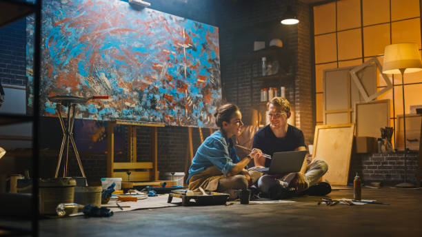 istock Couple of Artists Sitting in their Studio Use Laptop Computer, Working on a Project, Talking and Smiling. Painter and Her Partner Doing Research. Authentic Workshop with Oil Paintings and Tools 1183183588