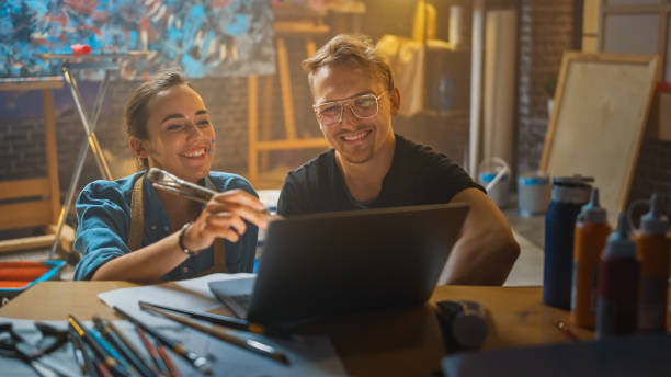 istock Couple of Artists Sitting in their Studio Use Laptop Computer, Working on a Project, Talking and Smiling. Painter and Her Partner Doing Research. Authentic Workshop with Oil Paintings and Tools 1183183616