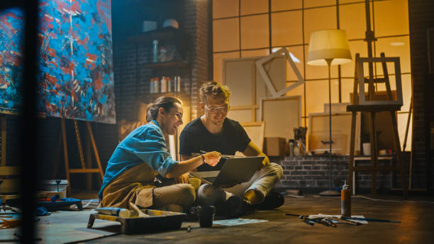 istock Couple of Artists Sitting in their Studio Use Laptop Computer, Working on a Project, Talking and Smiling. Painter and Her Partner Doing Research. Authentic Workshop with Oil Paintings and Tools 1183183633