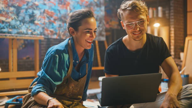 casal de artistas sentados em seu estúdio usar laptop computador, trabalhando em um projeto, falando e sorrindo. pintor e seu sócio que fazem a pesquisa. oficina autêntica com pinturas a óleo e ferramentas - painter paint art artist - fotografias e filmes do acervo