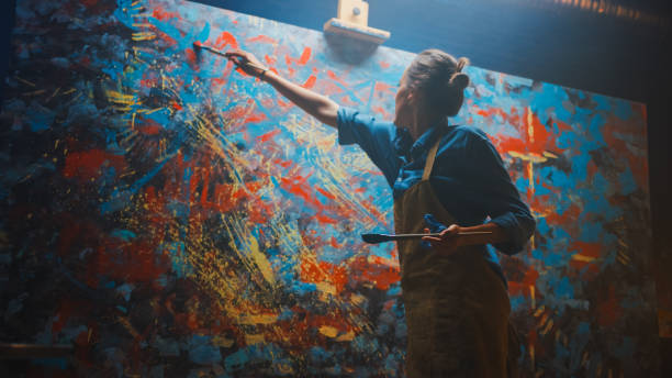 istock Talented Female Artist Energetically and Enthusiastically Using Paint Brush She Creates Modern Masterpiece of the Oil Painting. Creative Studio with Large Canvas of Striking Colors. Low Angle Shot 1183183668