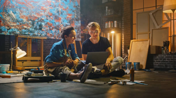 Couple of Artists Sitting in their Studio Use Laptop Computer, Working on a Project, Talking and Smiling. Painter and Her Partner Doing Research. Authentic Workshop with Oil Paintings and Tools