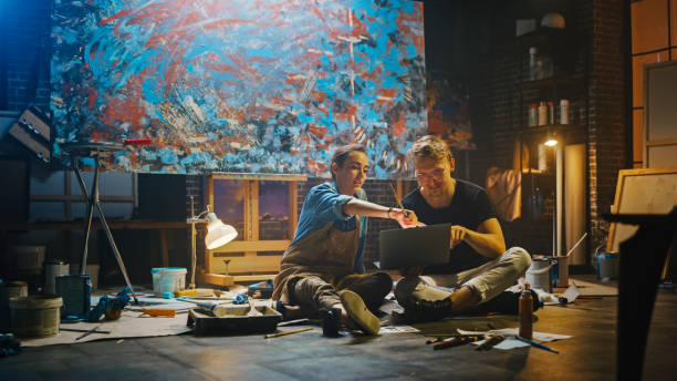 istock Couple of Artists Sitting in their Studio Use Laptop Computer, Working on a Project, Talking and Smiling. Painter and Her Partner Doing Research. Authentic Workshop with Oil Paintings and Tools 1183183717