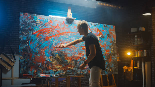 istock Talented Male Artist Energetically and Violently Using Paint Brush She Creates Modern Masterpiece of the Oil Painting. Dark and Messy Creative Studio with Large Canvas of Striking Colors 1183183733