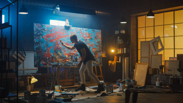 istock Talented Male Artist Energetically and Violently Using Paint Brush he Creates Modern Masterpiece of the Oil Painting. Dark and Messy Creative Studio with Large Canvas of Striking Colors 1183183769
