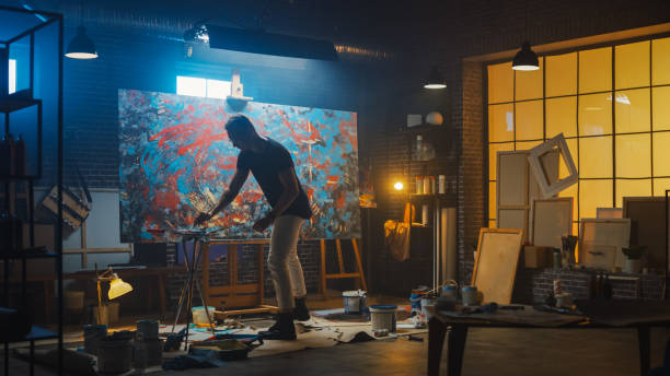 istock Talented Male Artist Energetically and Violently Using Paint Brush he Creates Modern Masterpiece of the Oil Painting. Dark and Messy Creative Studio with Large Canvas of Striking Colors 1183183780