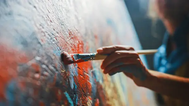 Photo of Female Artist Works on Abstract Oil Painting, Moving Paint Brush Energetically She Creates Modern Masterpiece. Dark Creative Studio where Large Canvas Stands on Easel Illuminated. Low Angle Close-up