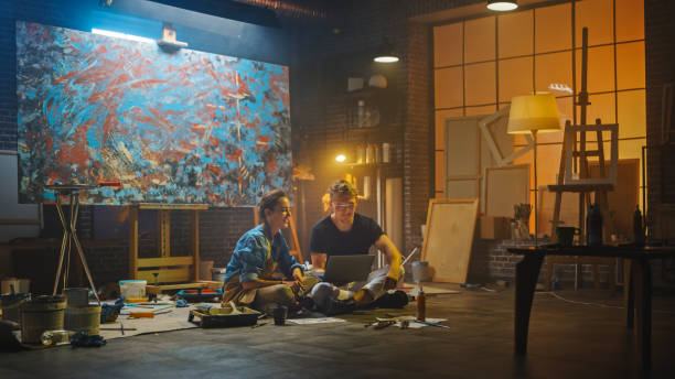 istock Couple of Artists Sitting in their Studio Use Laptop Computer, Working on a Project, Talking and Smiling. Painter and Her Partner Doing Research. Authentic Workshop with Oil Paintings and Tools 1183183785