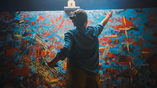 Talentosa artista femenina trabaja en pintura al óleo abstracta, usando pincel que crea obra maestra moderna. Estudio creativo oscuro y desordenado donde Grandes lienzos se para nado sobre Easel Iluminado. photo