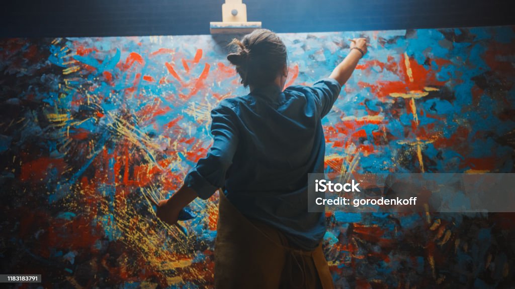 Talented Female Artist Works on Abstract Oil Painting, Using Paint Brush She Creates Modern Masterpiece. Dark and Messy Creative Studio where Large Canvas Stands on Easel Illuminated. Artist Stock Photo
