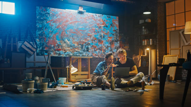 istock Couple of Artists Sitting in their Studio Use Laptop Computer, Working on a Project, Talking and Smiling. Painter and Her Partner Doing Research. Authentic Workshop with Oil Paintings and Tools 1183183800