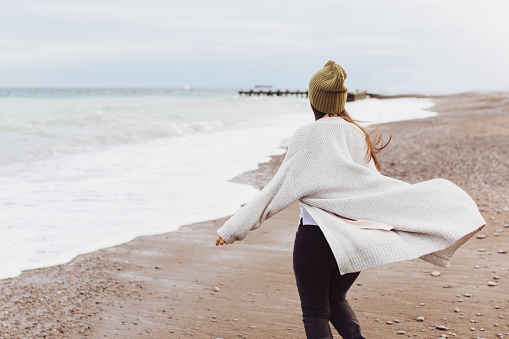A beautiful young girl walks along the seashore, a storm, hair fly apart, a gray cardigan, a sports figure, in sneakers, enjoys, autumn or winter