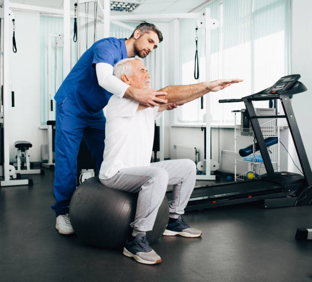 patient aîné ayant rendez-vous de thérapie physique avec son docteur. homme faisant l'exercice de poteau de réadaptation tout en s'asseyant sur une bille de forme physique. - centre de désintoxication photos et images de collection