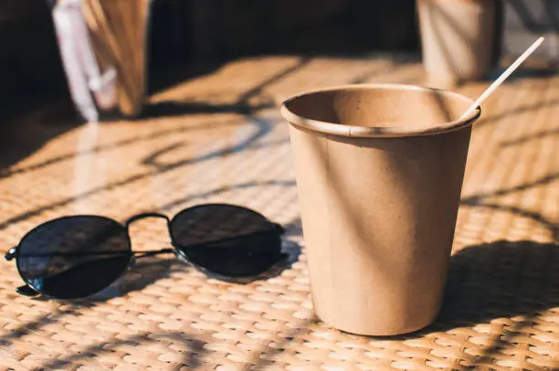 Disposable paper coffee cup on table in cafe with wooden stirrer,near sunglasses.Environmentally friendly lifestyle.Zero Waste,Save The Planet,Earth Day,No Plastic,Recycling Concept