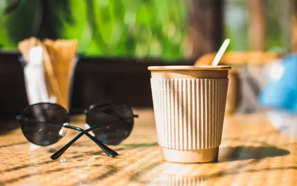 Disposable paper coffee cup with holder on table in cafe with wooden stirrer,near sunglasses.Environmentally friendly lifestyle.Zero Waste,Save The Planet,Earth Day,No Plastic,Recycling Concept