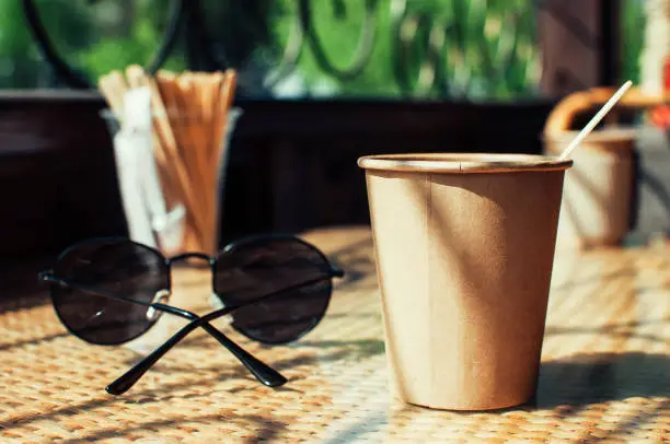 Disposable paper coffee cup on table in cafe with wooden stirrer,near sunglasses.Environmentally friendly lifestyle.Zero Waste,Save The Planet,Earth Day,No Plastic,Recycling Concept