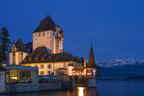 nightscape do castelo de oberhofen que olha para fora lago thun e iluminado no crepúsculo. montanhas cobertas de neve e céu pitoresco no fundo. cantão de berna. - lake thun switzerland night lake - fotografias e filmes do acervo