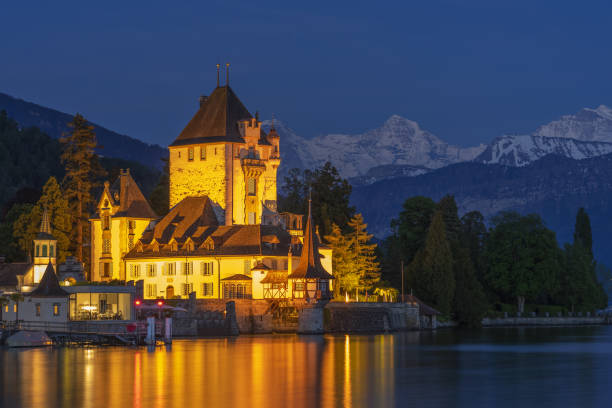 nightscape do castelo de oberhofen que olha para fora lago thun e iluminado no crepúsculo. montanhas cobertas de neve e céu pitoresco no fundo. cantão de berna. - lake thun switzerland night lake - fotografias e filmes do acervo