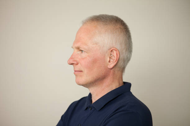 retrato de estudio de cerca de un hombre de pelo gris de 55 años con un polo azul sobre un fondo beige - mejillas enrojecidas fotografías e imágenes de stock