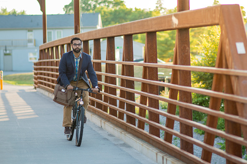An attractive Indian businessman is bicycling to work. He is on a bridge on a sunny, summer day. He smiles widely as his suit jacket and bag blow in the wind.