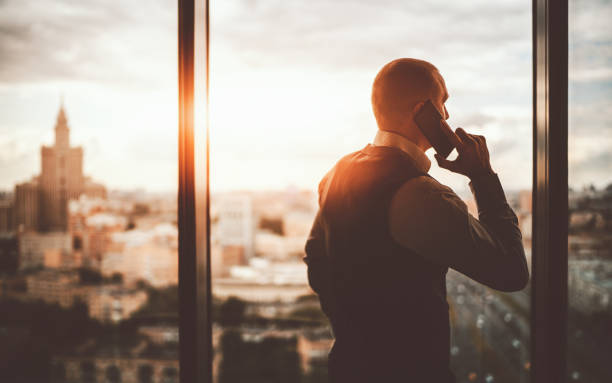 Businessman phoning near the window Silhouette of a prosperous man entrepreneur standing near the panoramic window of a luxurious business office skyscraper and pensively looking on an evening cityscape while talking on the phone wealthy stock pictures, royalty-free photos & images