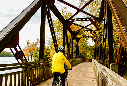Senior male bicyclist in yellow jersey riding old railroad bridge converted into recreation trail; Missouri River on the left; autumn in Missouri