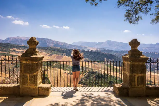 An Asian woman in her 40s is enjoying the spectacular view of the mountains and valley. She's using her mobile phone to take a photo while on her Summer vacation. Ronda is a mountaintop city in Spain’s Malaga province that’s set dramatically above a deep gorge.