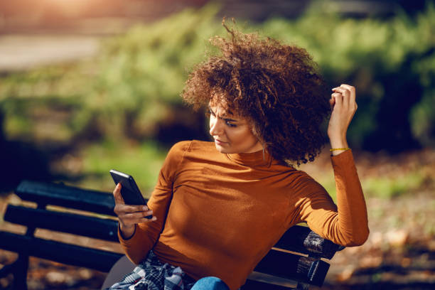 serious mixed race young woman with curly hair and in turtleneck sitting in park on bench and using smart phone for reading or writing message. - using phone garden bench imagens e fotografias de stock