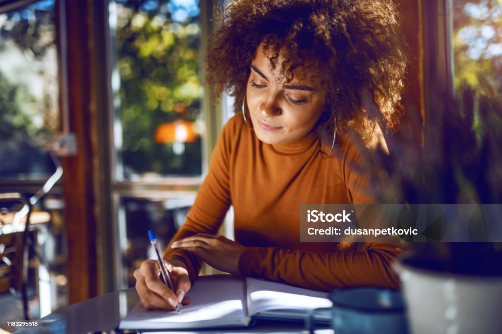 Portrait of cute mixed race student with curly hair and in turtleneck sitting in cafe and studying for exams. Writing - Activity Stock Photo