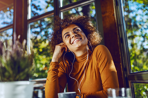 Portrait of attractive smiling mixed race young woman sitting in cafe and listening music and looking away.