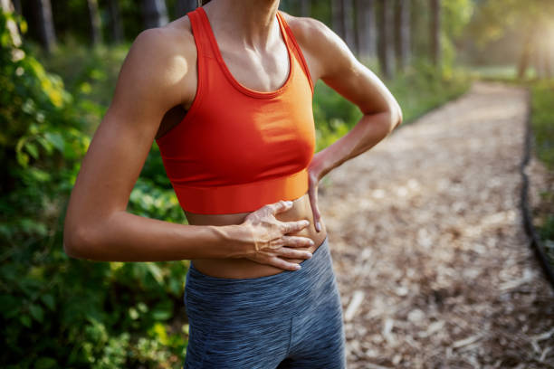 abgeschnittenes bild von sitzen schlanken sportlichen kaukasischen frau in sportbekleidung auf dem weg in wäldern stehen und halten magen. sonniger tag in der natur. - belly ache stock-fotos und bilder