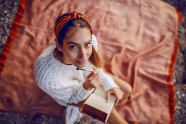 vista dall'alto dell'attraente bruna caucasica in maglione e con fascia seduta su coperta all'aperto e tenendo penna e diario in mano mentre si guarda la fotocamera. - beach ideas foto e immagini stock