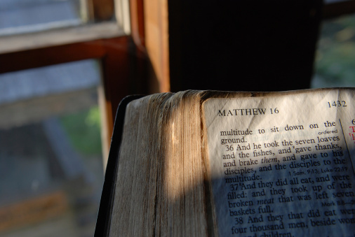 Sunlight shines through the window of a home, illuminating a page of an old Bible turned to the Gospel of Matthew, chapter 16.