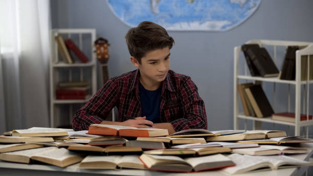 istock Schoolboy looking at huge pile of open books on table doing homework, education 1183307374