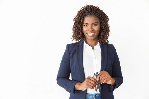 Happy successful manager holding glasses. Young African American business woman standing isolated over white background, looking at camera, smiling. Glasses wearer concept