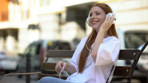 Excited caucasian teen girl in headset listening to favorite song, resting bench