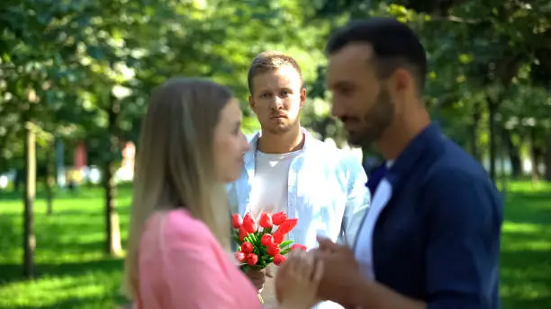 Disappointed man with flowers looking at girlfriend flirting with another man