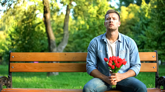Waiting handsome man with red flowers sitting park bench, outdoor date affection