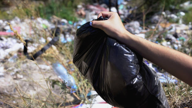 istock Womans hand holding bag with waste against landfill in forest, ecological system 1183329667