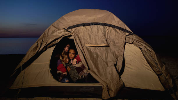 Three people sit closely together inside a tent at dusk, looking out with serious expressions under a darkening sky.