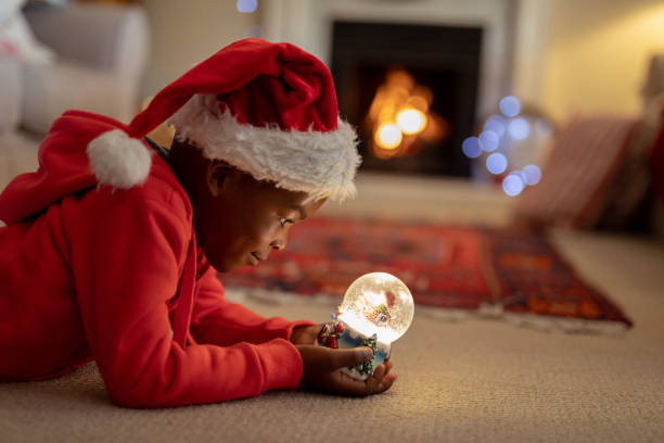 Boy at home at Christmas time Side view of a young mixed race boy in his sitting room at Christmas, wearing a santa hat and holding a snow globe snow globe photos stock pictures, royalty-free photos & images