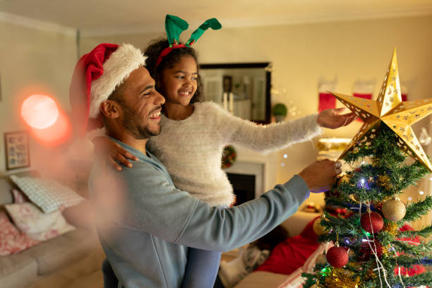 Family at home at Christmas time Side view of a mixed race man with his young daughter in their sitting room at Christmas, wearing festive hats, the man holding his daughter and touching a Christmas tree traditional sport stock pictures, royalty-free photos & images