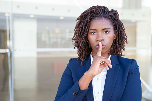 Serious silent professional keeping secret. Young black business woman standing at outdoor glass wall, applying index finger at mouth, making shh gesture. Conspiracy concept