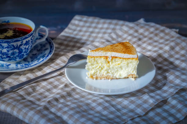 istock On a dark countertop, on a light checkered linen napkin, a white and blue patterned tea pair with tea and saucer with a piece of classic cheesecake and a fork. 1183357212