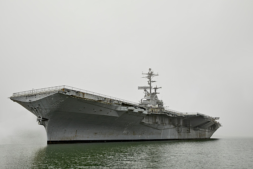 A Vintage WWII era aircraft carrier military ship lies still in the water surrounded by heavy fog.