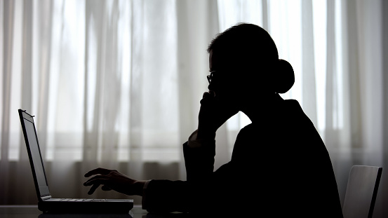 Dark silhouette of businesswoman talking on phone at night making note on laptop