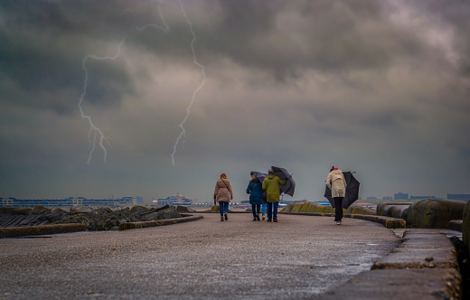 Ijmuiden/Holland - October 06 2019: A group of people walking and trying to keep umbrellas in a windy day and lightning in background